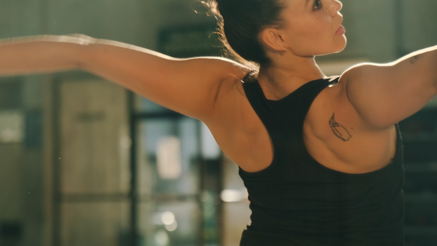 A female athlete extends her arms wide during a workout session in a gym, showing a shoulder tattoo.