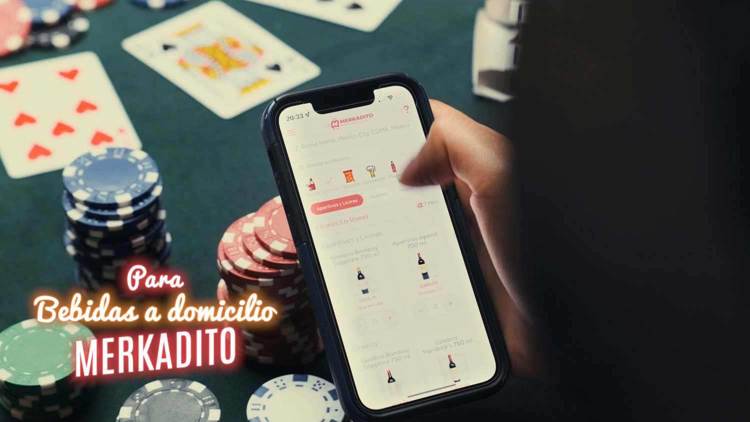 Hand holding smartphone showing Merkadito app for bebidas a domicilio near poker chips and cards.