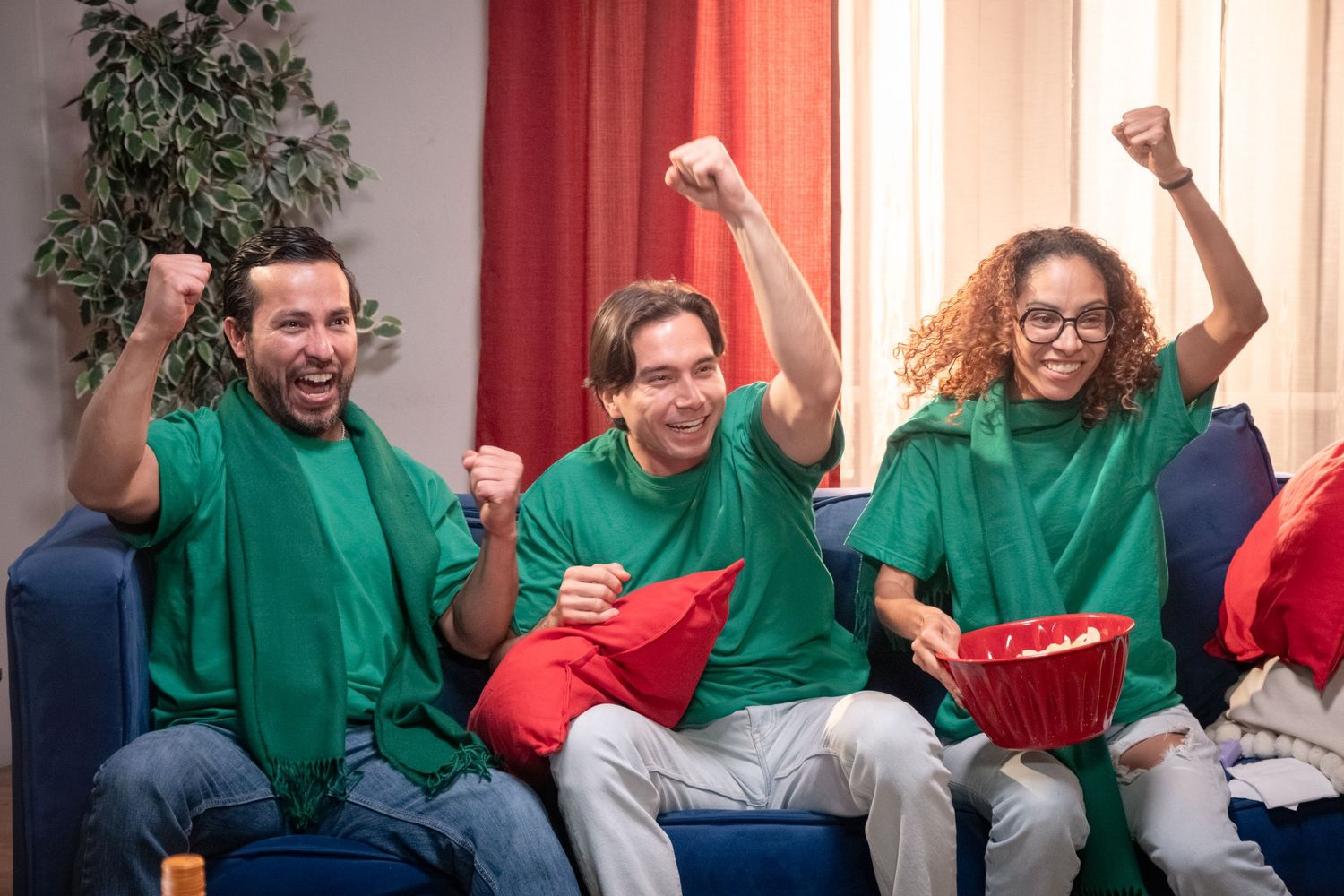 Three friends in green shirts celebrate together on a sofa with popcorn while watching sports.
