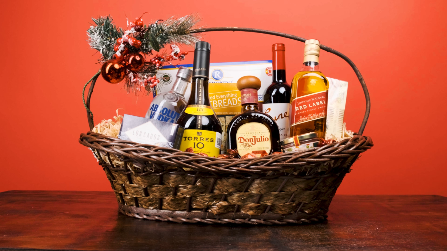 Wicker gift basket filled with whiskey bottles and gourmet items on red holiday background.