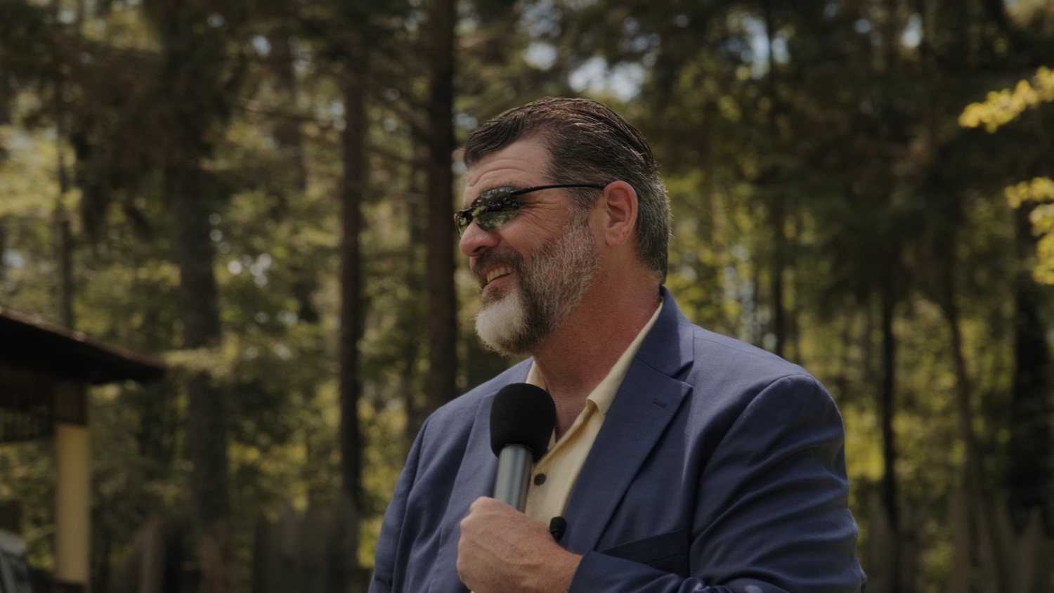 Bearded man in blue blazer speaking into microphone outdoors among trees.