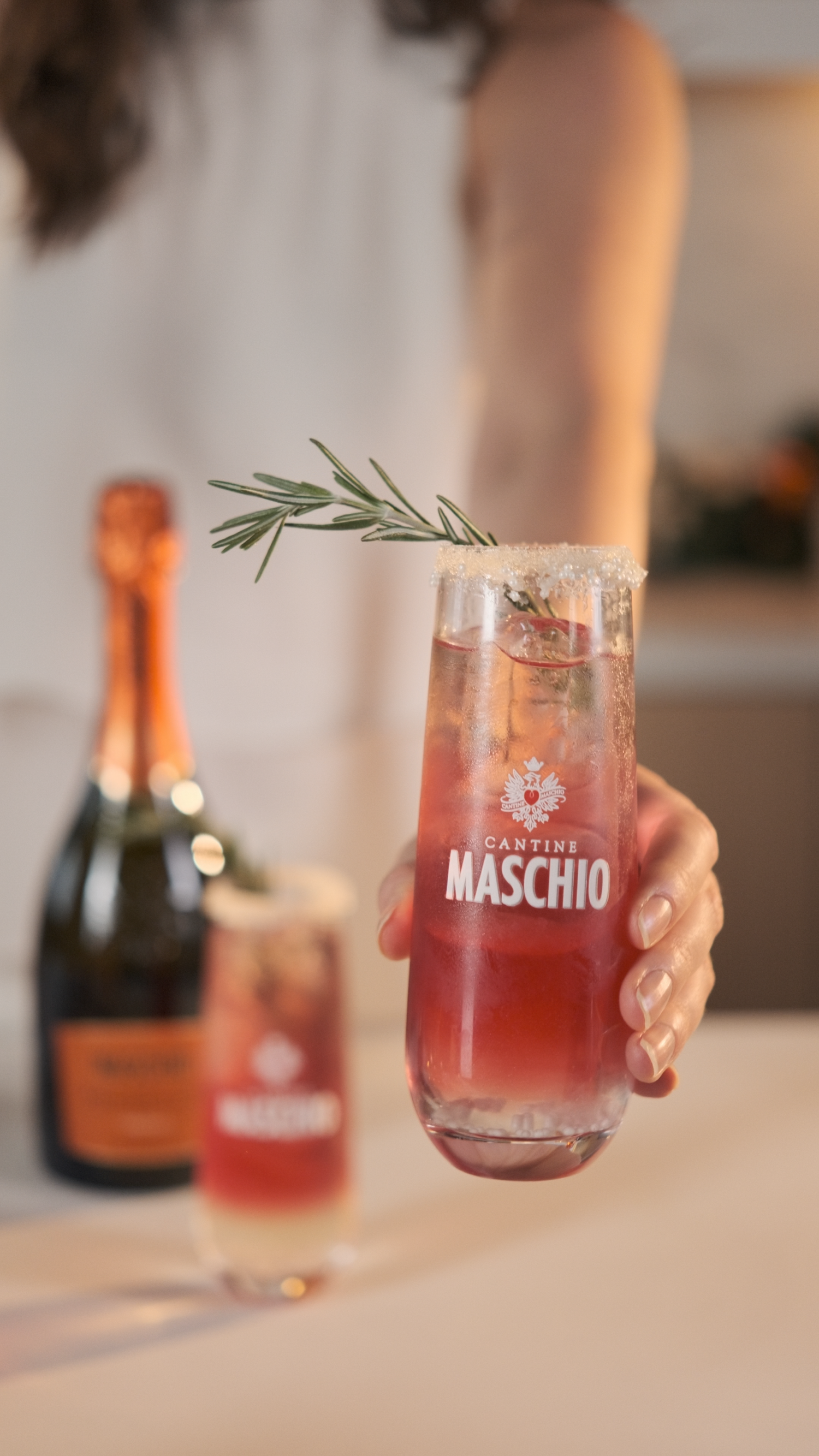 Cantine Maschio branded cocktail glass with rosemary sprig held up in kitchen setting.