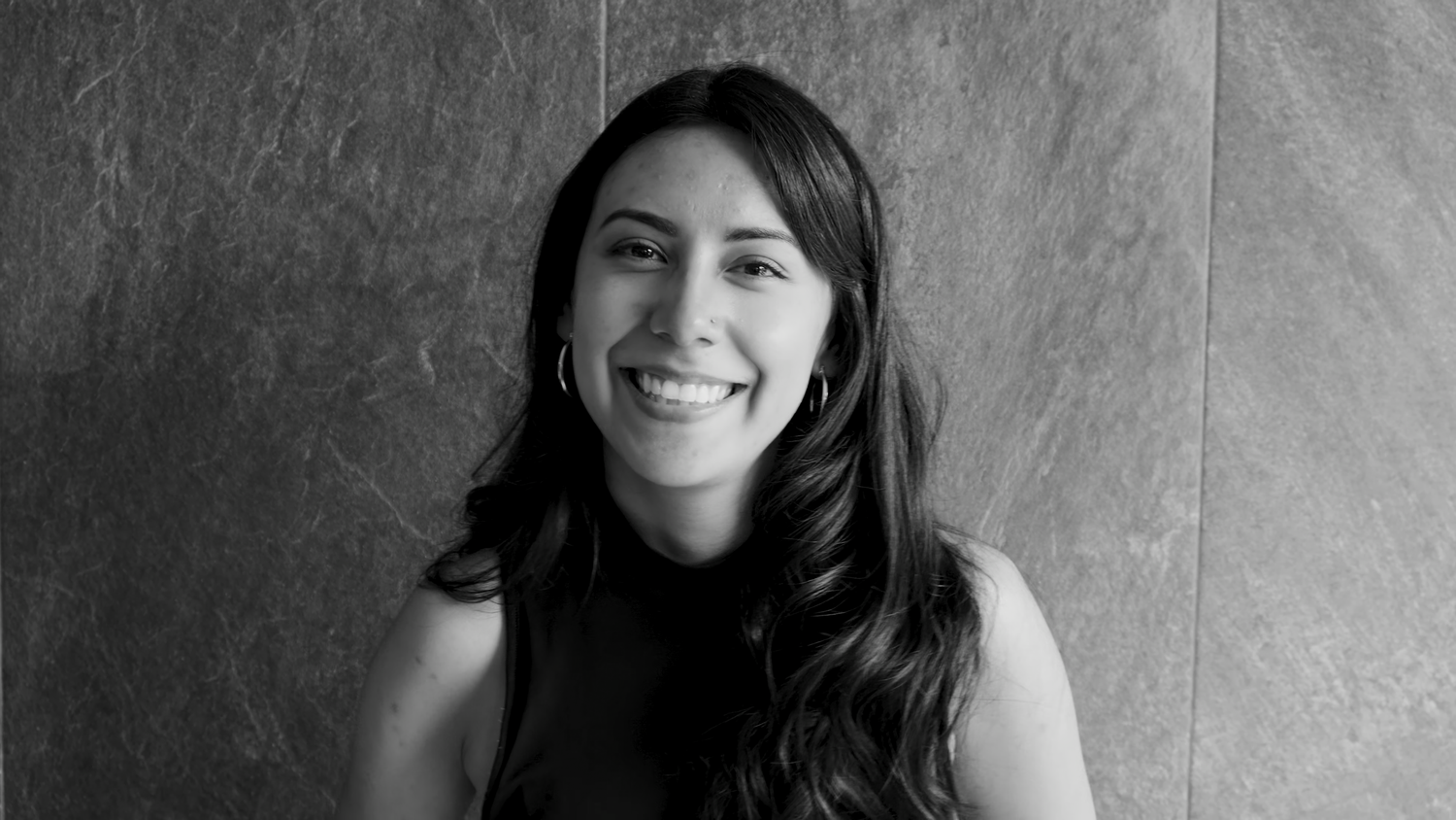 Black and white portrait of smiling woman with long wavy hair against stone wall.