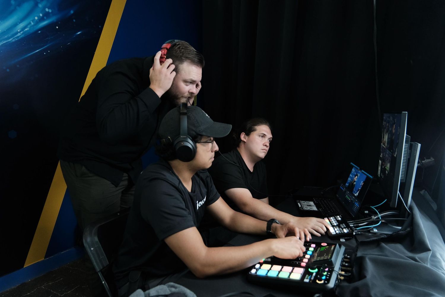 Three production crew members in black operate live streaming equipment and mixing boards at a conference event.
