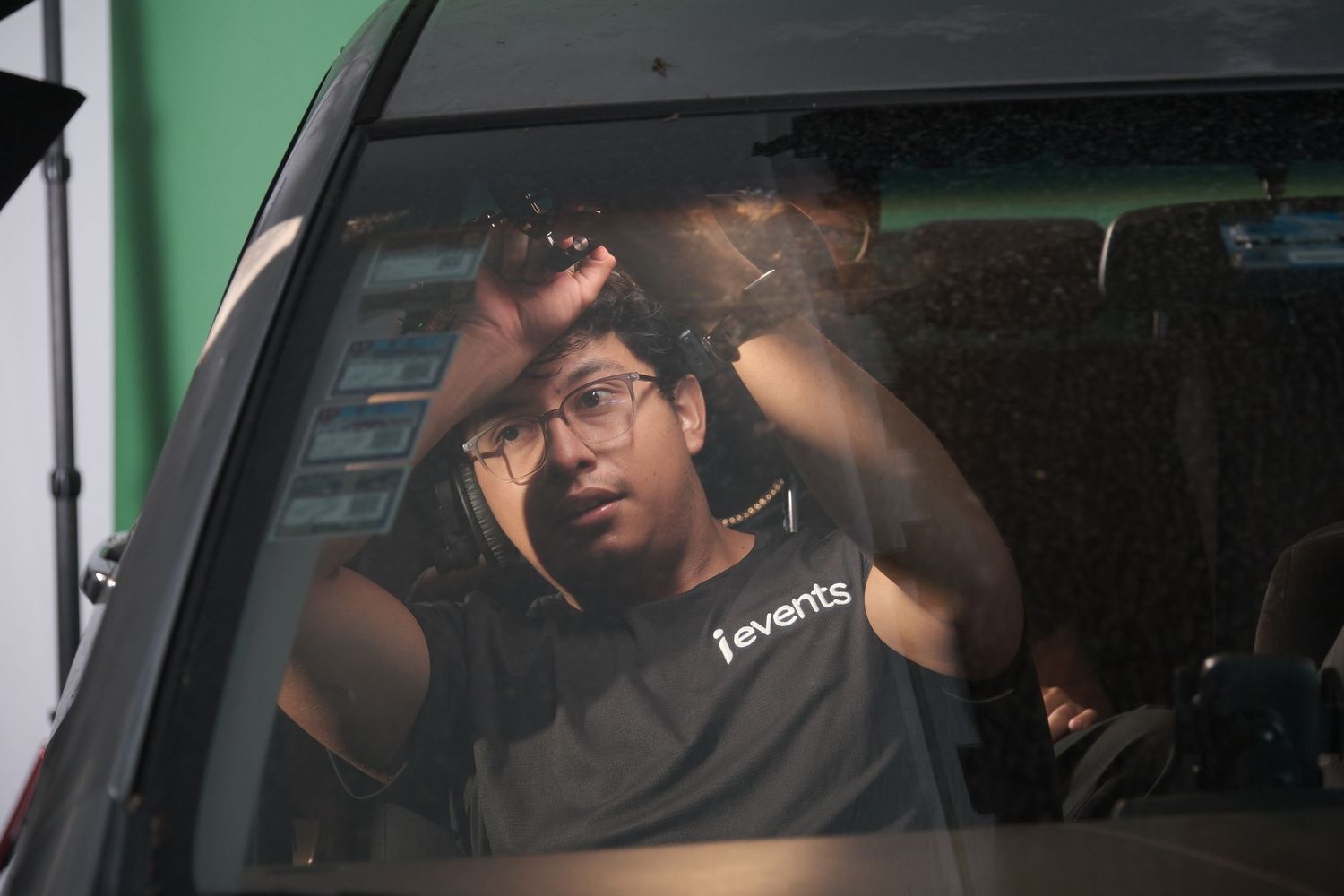 Young man in events staff shirt sits in car, adjusting hair while looking in rearview mirror.