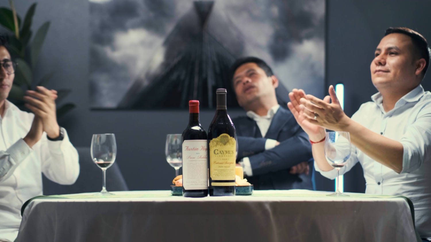 Three men applauding at a dining table with wine bottles and glasses in a restaurant.