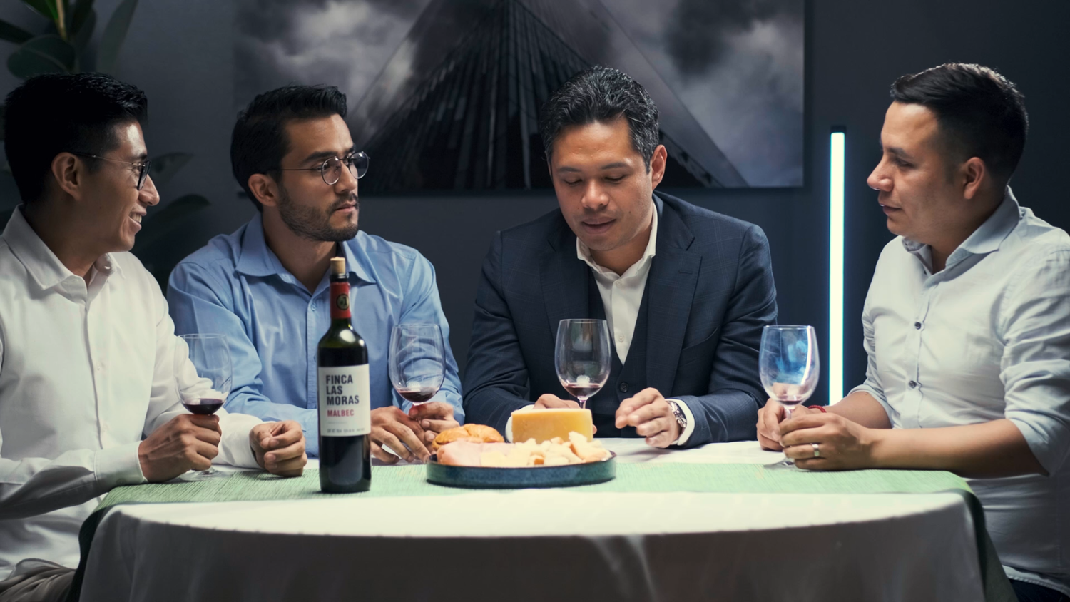 Four men sit at a dinner table with wine glasses and a bottle of red wine in a dimly lit restaurant.