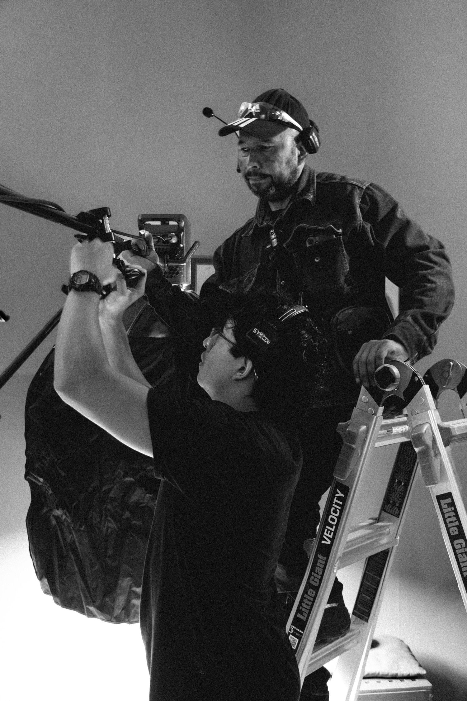 Black and white BTS photo of two filmmakers adjusting camera equipment on a ladder during production.