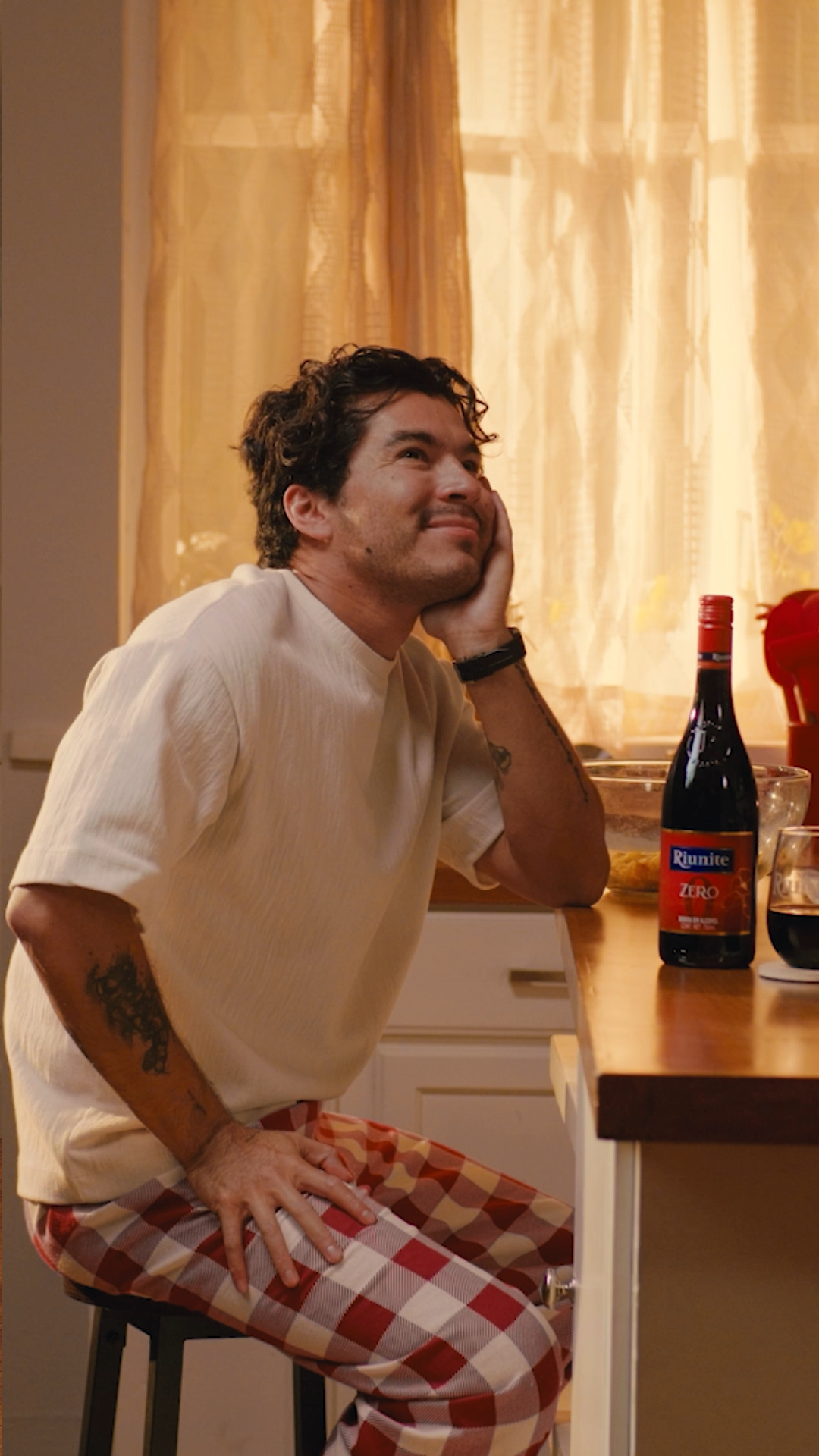 Smiling young man in pajama pants sitting at a kitchen counter with wine bottles nearby.