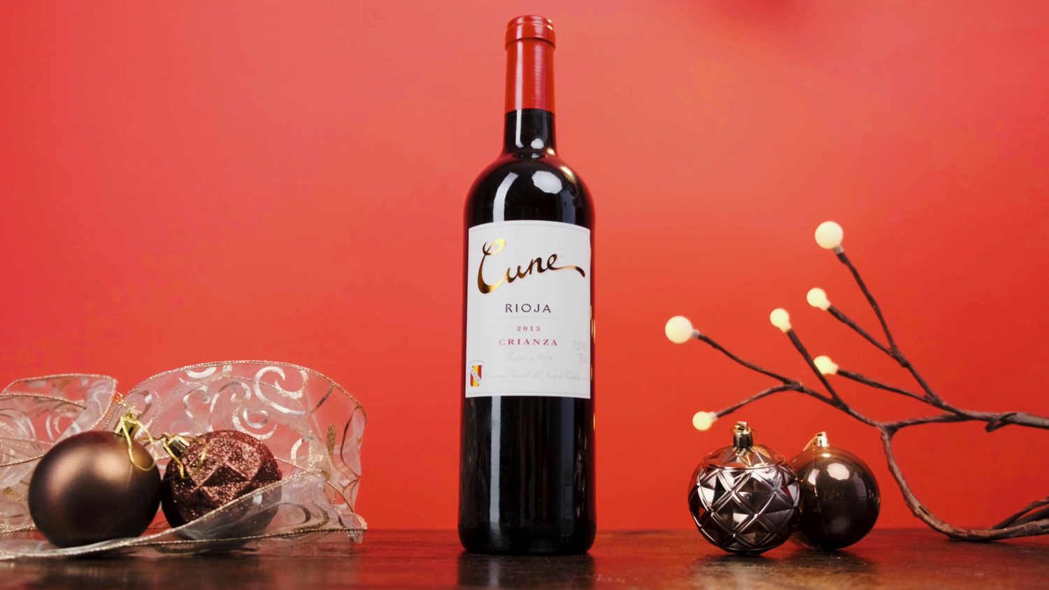 Cune Rioja Reserva red wine bottle surrounded by dark holiday ornaments on a festive red backdrop.