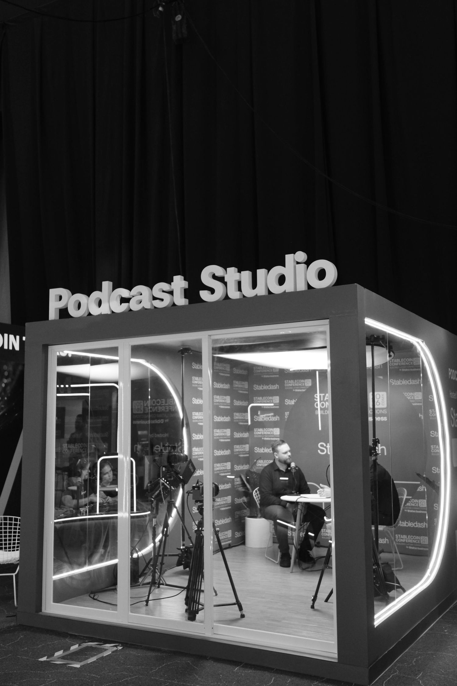 Black and white exterior view of a glass-enclosed Podcast Studio booth at a professional conference event.