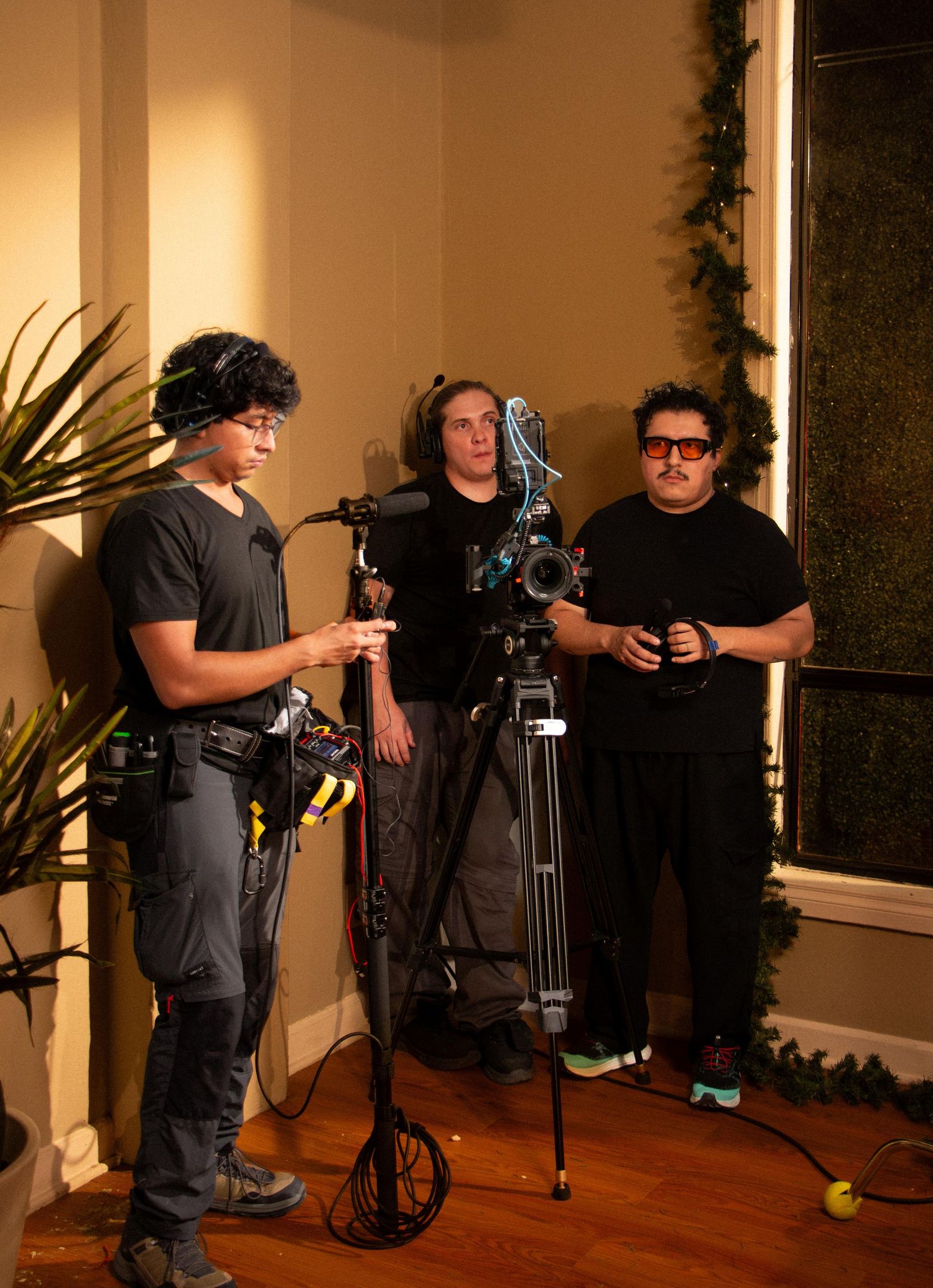 Three film crew members with camera equipment standing near a window on an indoor movie set.