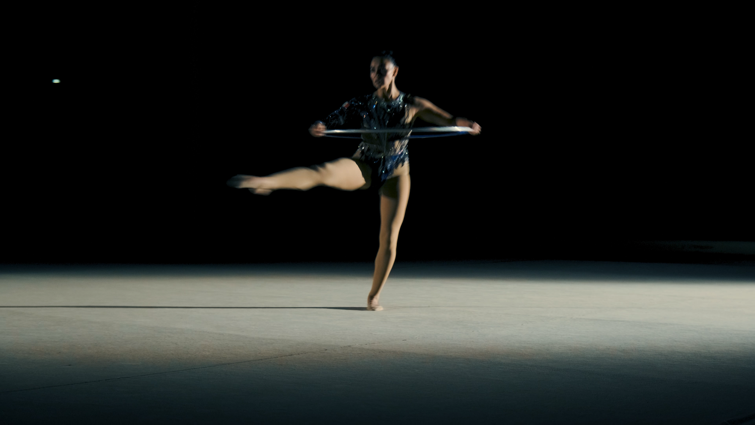 Rhythmic gymnast balances on one leg while spinning hoop in spotlight on dark arena floor.
