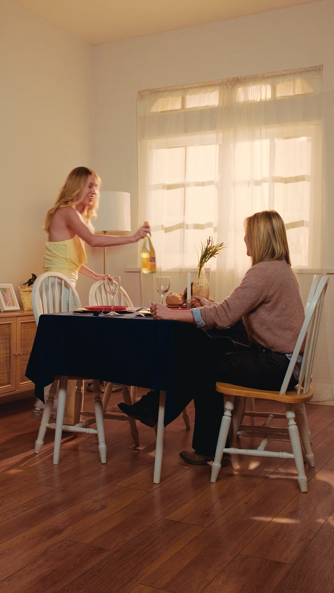 Blonde woman in yellow top presenting a wine bottle to a seated woman at dinner table.