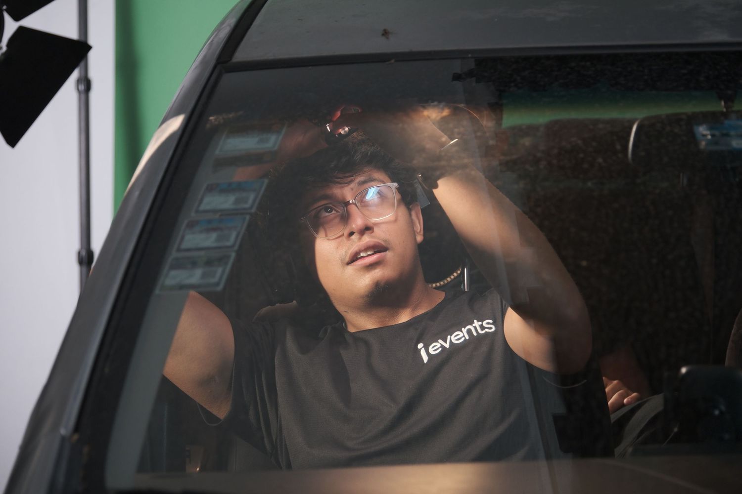 A film crew member in a black shirt adjusting equipment inside a vehicle on a green screen set.
