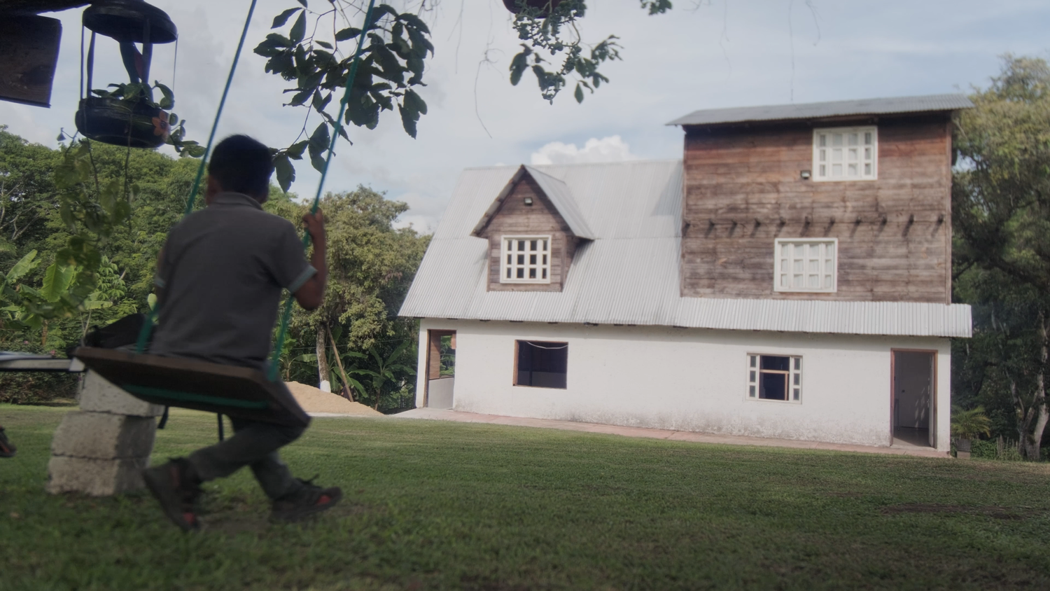 Person sitting on a swing in a lush garden with a rustic wooden two-story house in the background.