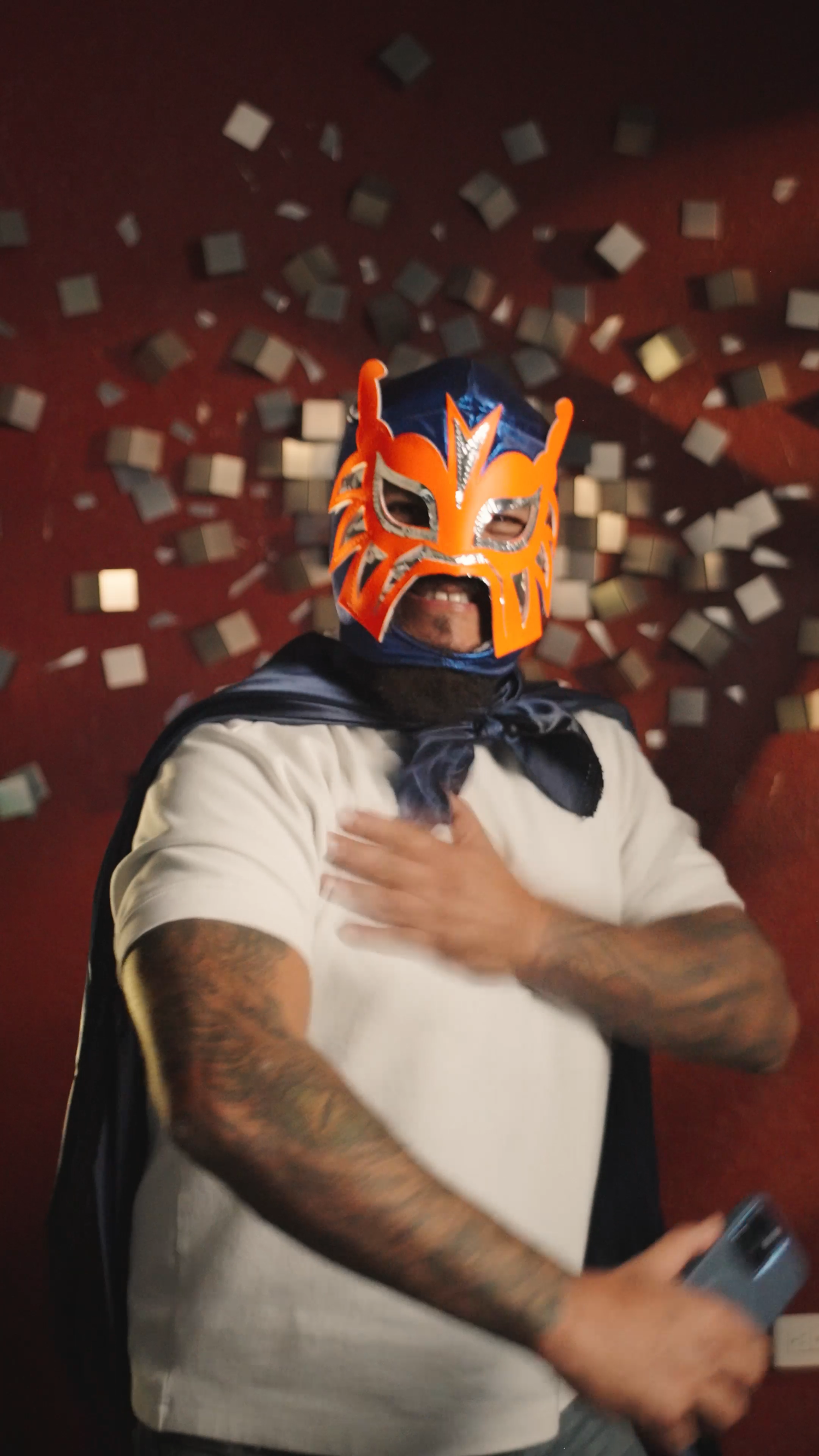 A wrestler wearing a colorful blue and orange lucha libre mask and cape poses dramatically against a red wall.