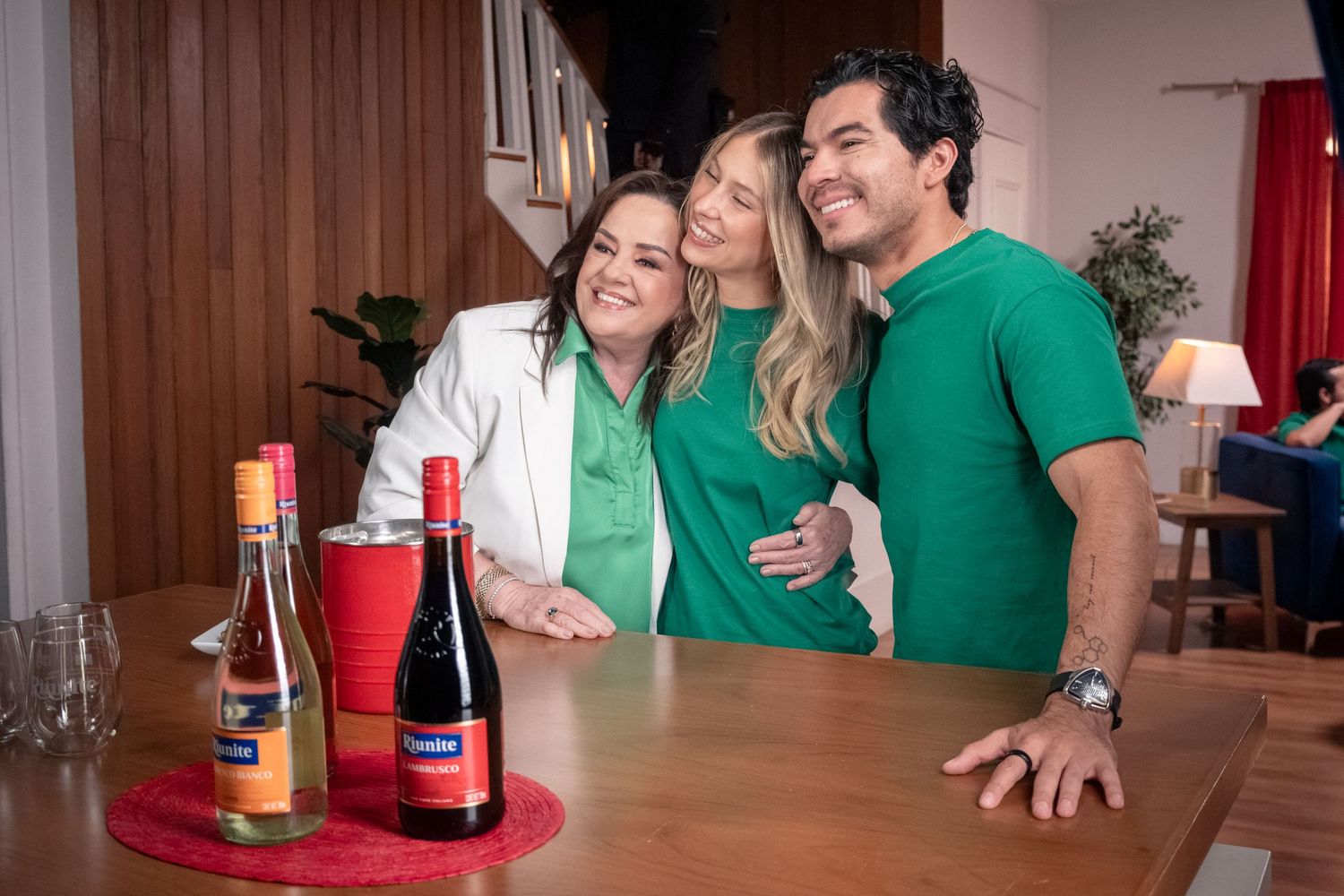 Three people smiling together behind a table with Riunite wine bottles in a cozy living room.