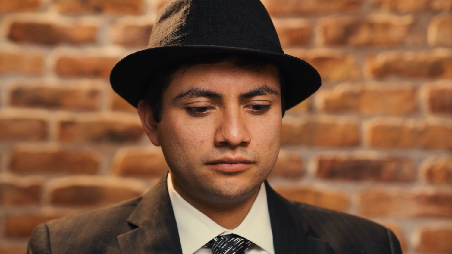 Young man wearing a black fedora hat and suit looking down against a brick wall background.