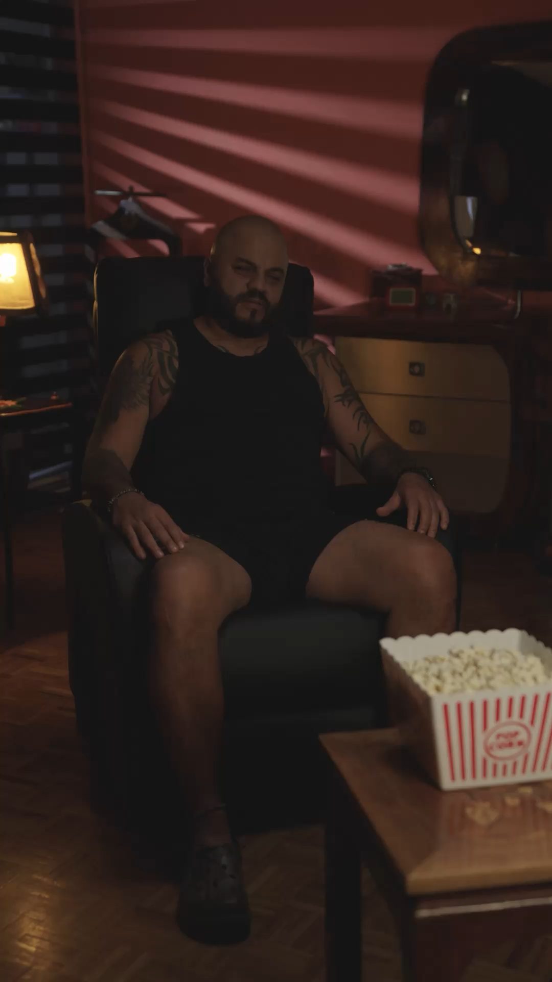 Tattooed bald man sitting in an armchair in a dimly lit red room with a bucket of popcorn nearby.