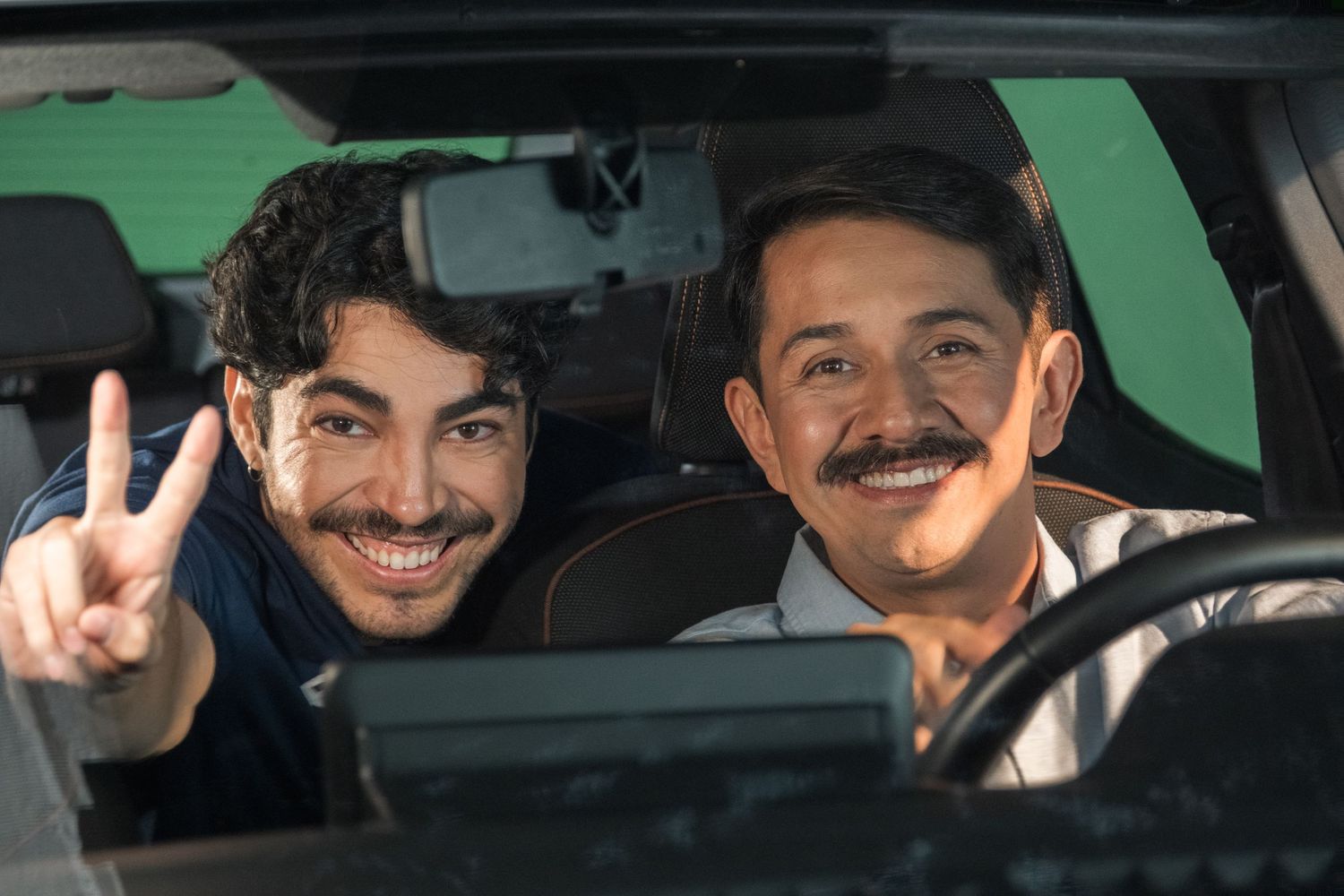 Two smiling men posing inside a car on a green screen film set.