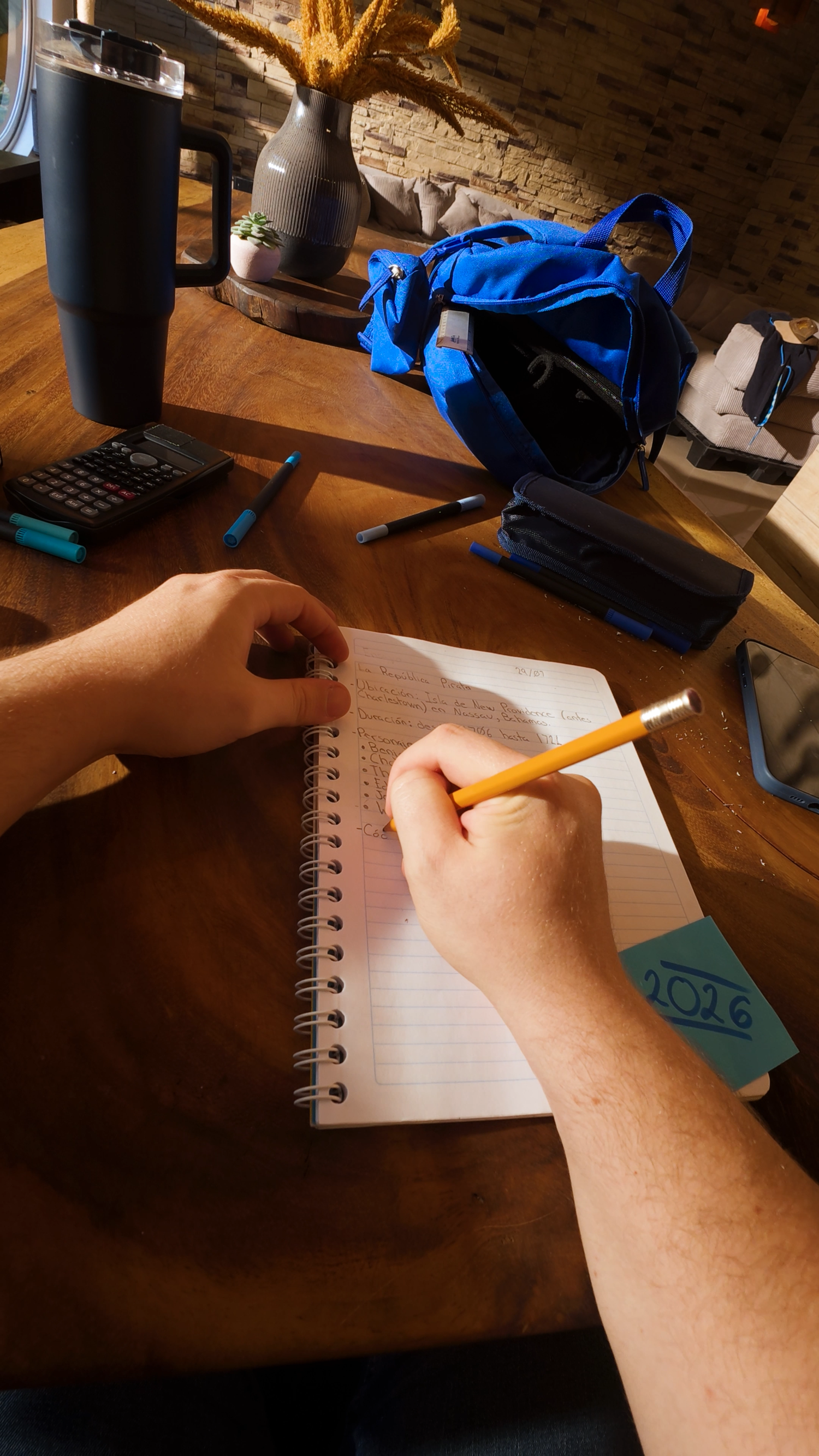 Close-up of hands writing in spiral notebook with pencil on wooden desk with backpack.