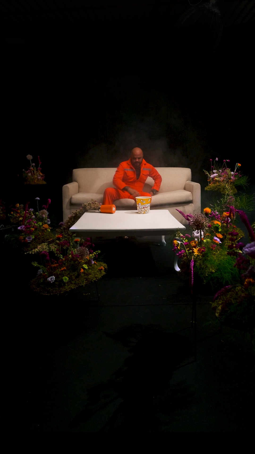 Man in orange jumpsuit sitting on beige sofa surrounded by colorful flowers in dark studio setting.