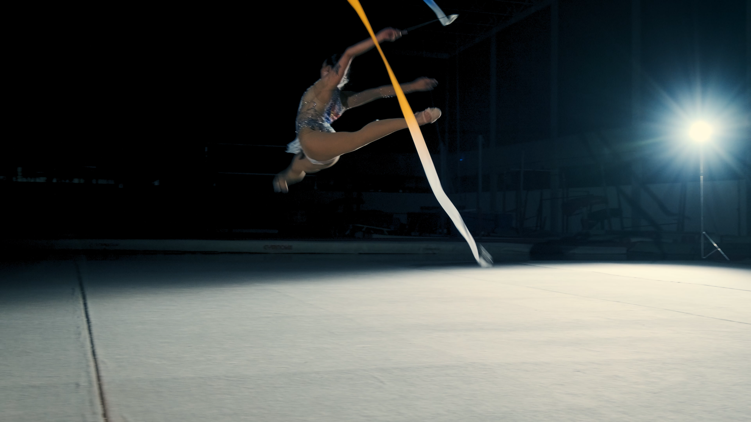 Rhythmic gymnast leaps high in air with ribbon during stunning athletic performance in dark arena.