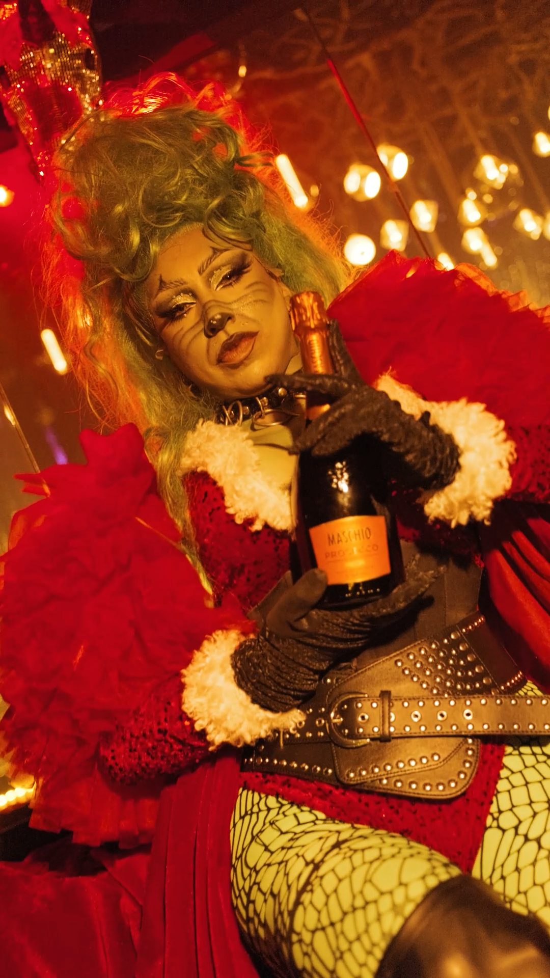 Drag performer dressed as Grinch in red costume holding Veuve Clicquot champagne bottle.