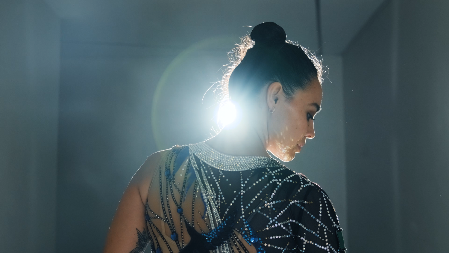 Gymnast in a sparkling leotard with hair in a bun posing under a bright backlight.