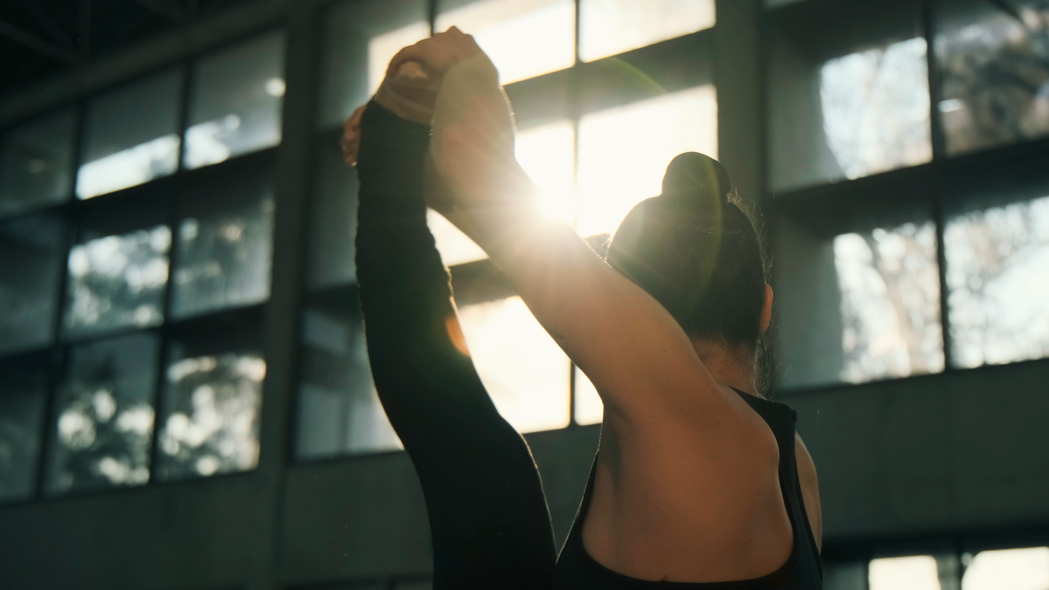 A female athlete stretches her arms upward in a sunlit indoor sports facility.