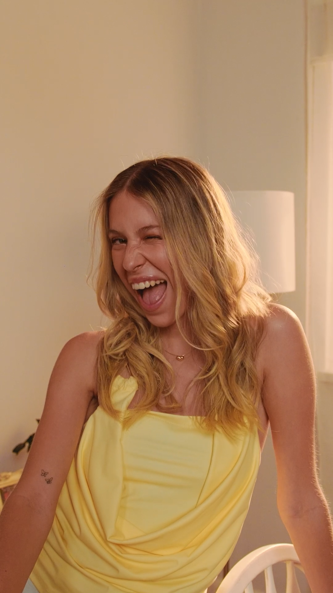 Laughing blonde woman in yellow dress with wavy hair in a bright cheerful setting.