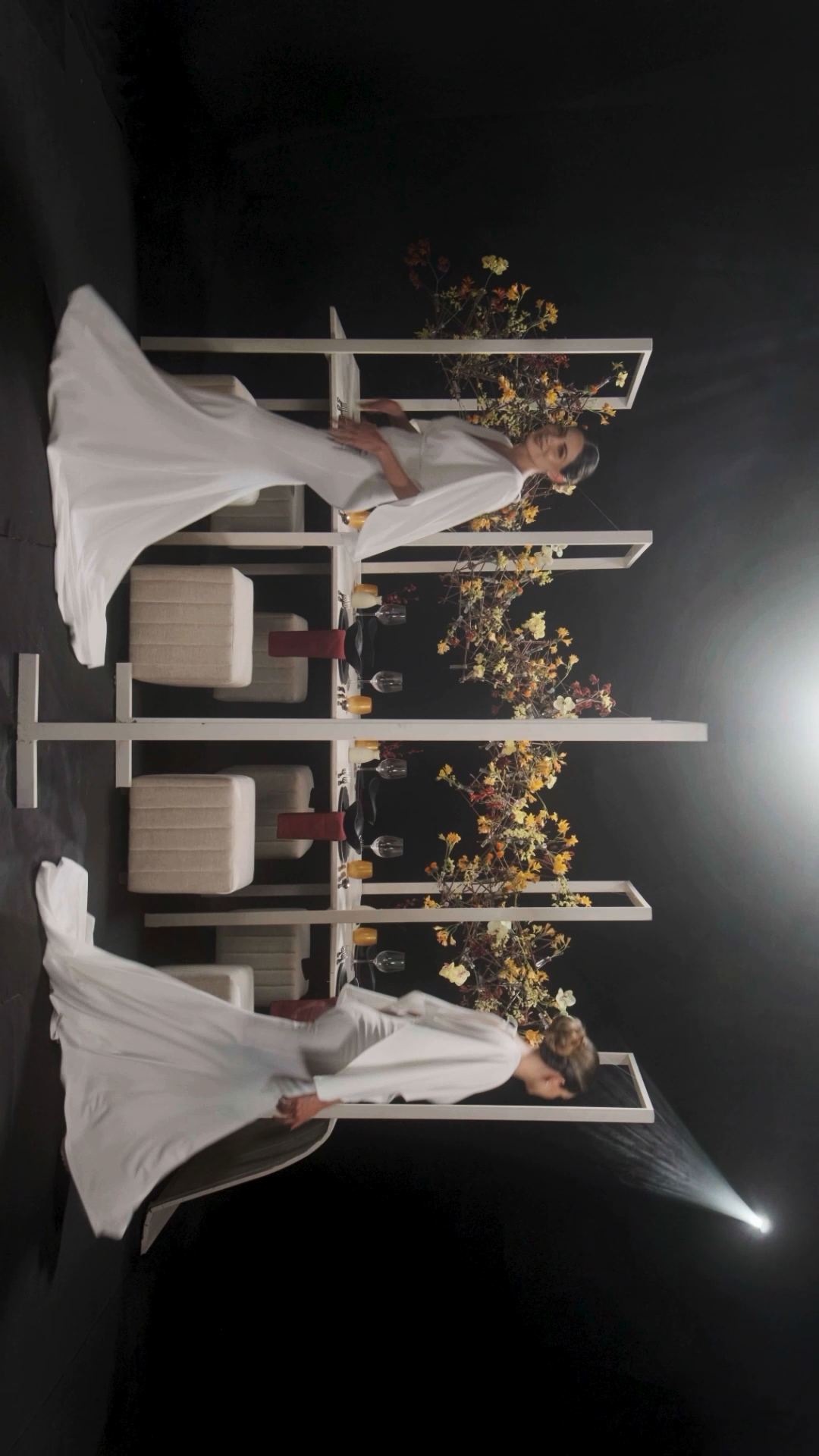 Two women in flowing white gowns pose symmetrically beside white geometric frames with yellow floral arrangements.