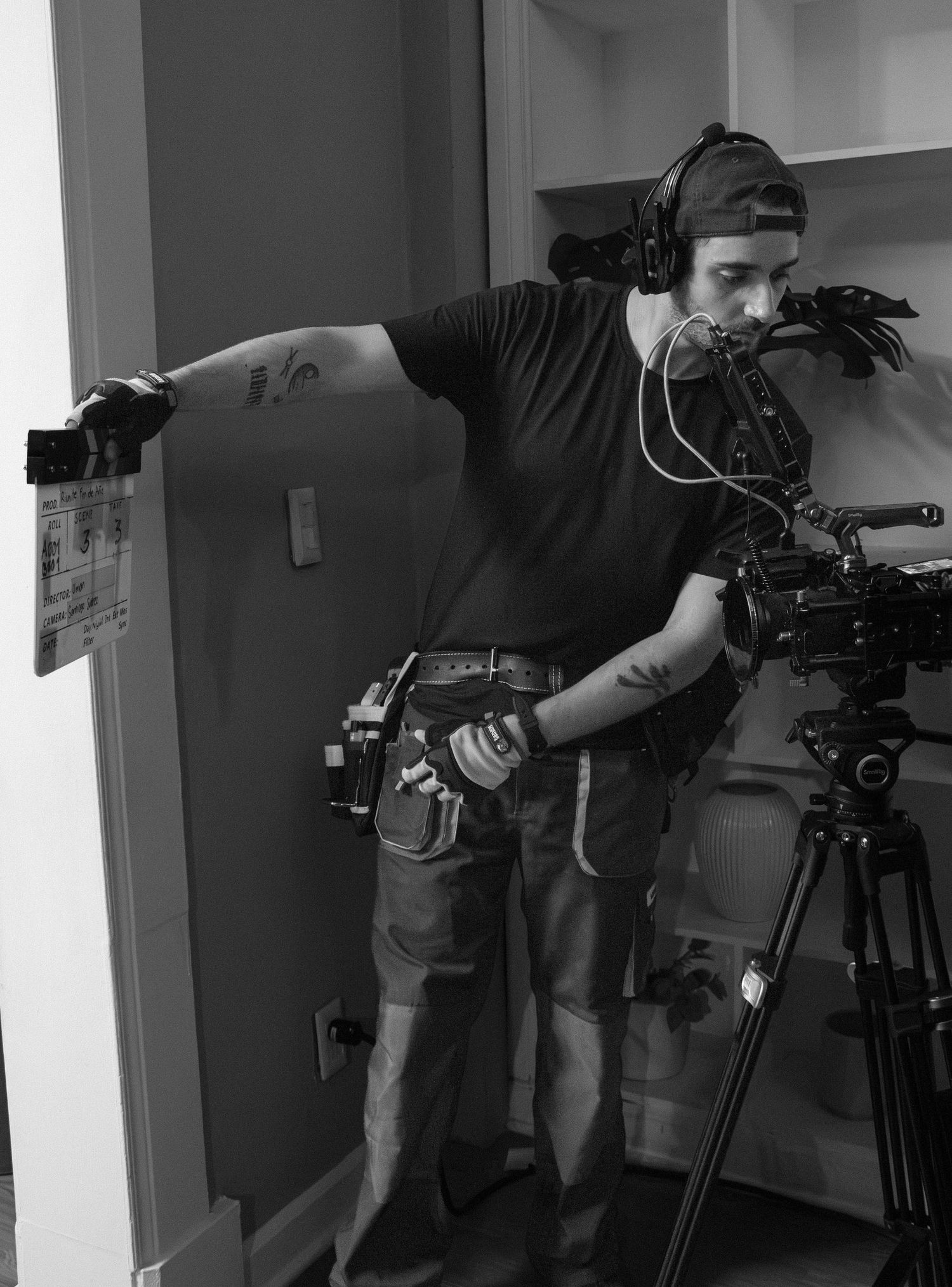 Black and white photo of film crew member holding clapperboard next to professional camera on tripod.