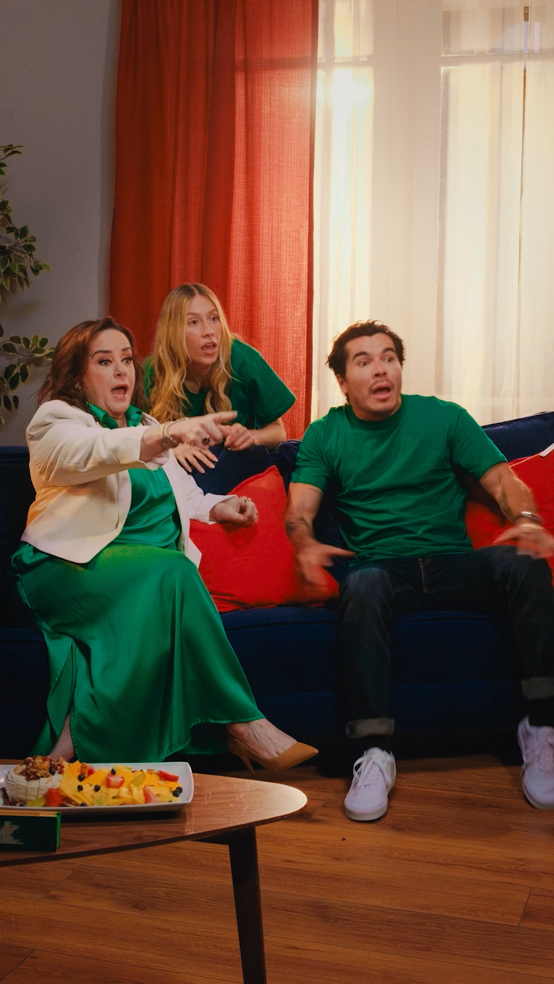 Three excited friends react emotionally while watching TV together on a blue sofa in a warm living room.