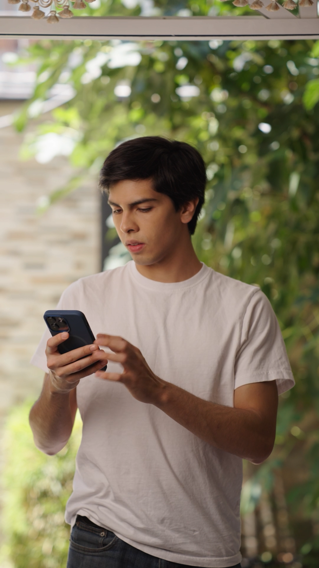 Young man in white t-shirt using smartphone outdoors with green foliage background.