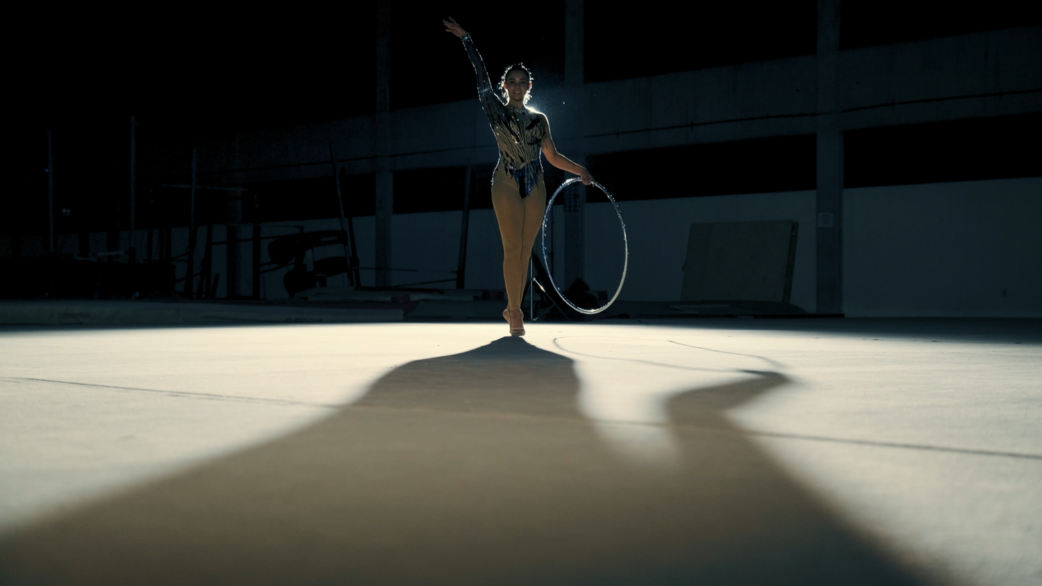 Rhythmic gymnast performing hoop routine under dramatic spotlight in dark arena.