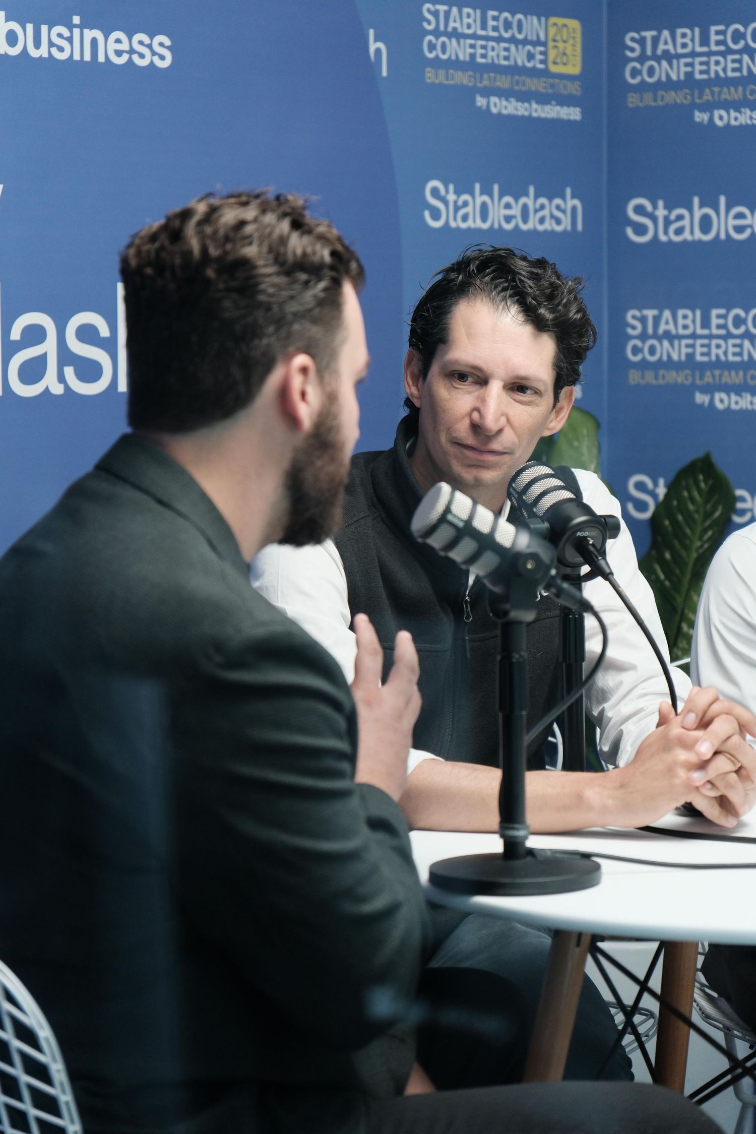 Two men engage in conversation at a podcast recording table with microphones at the Stabledash Stablecoin Conference.