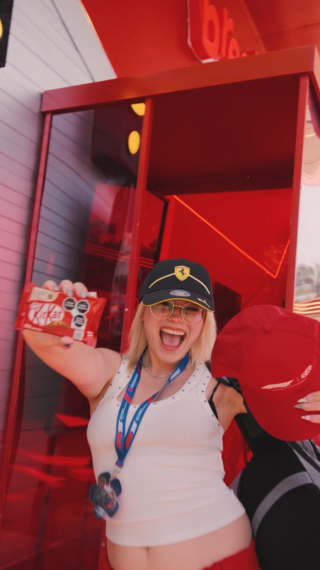 Excited blonde woman holding KitKat package at a red Ferrari-themed promotional event outdoors.
