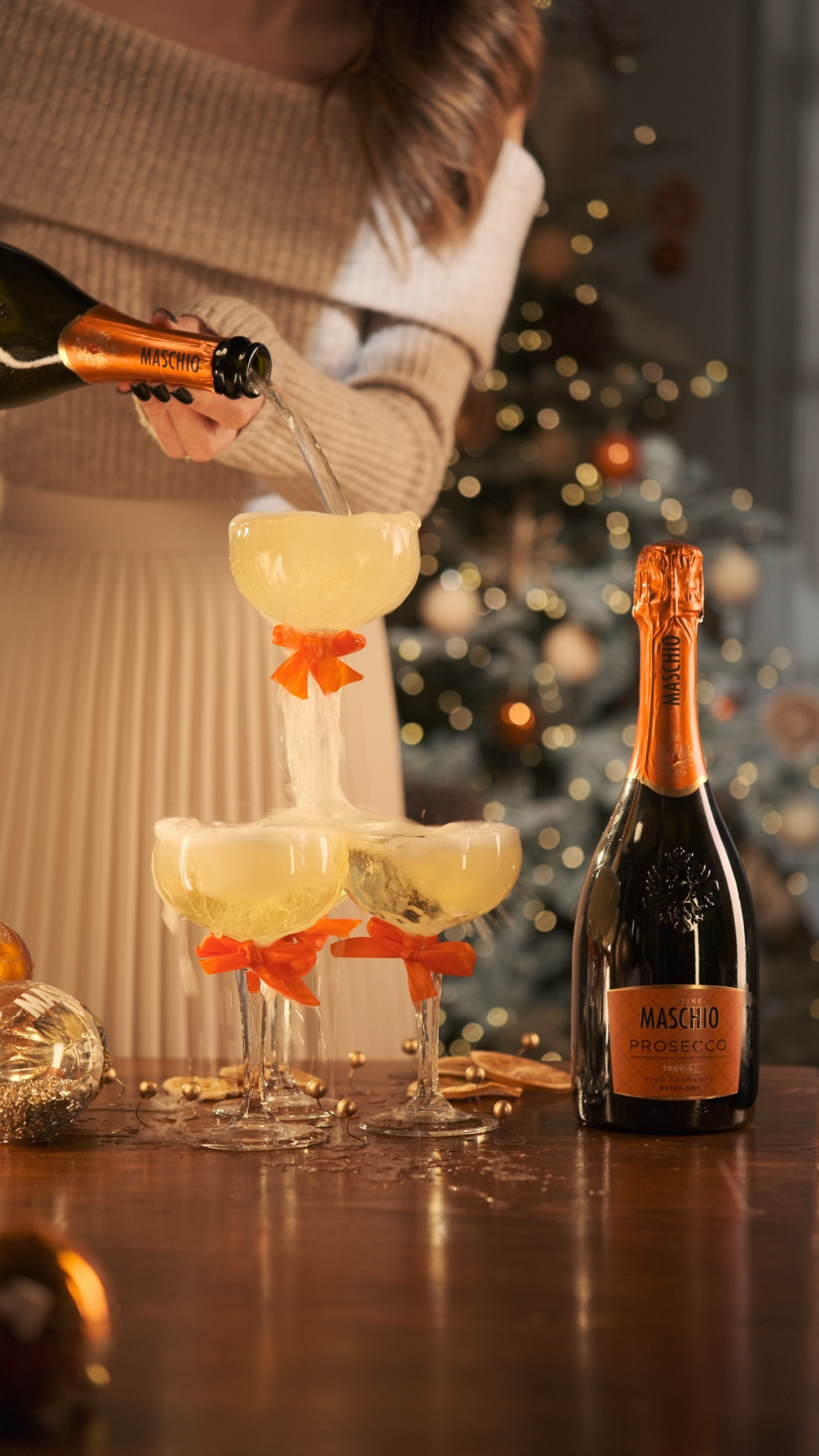Woman pouring Maschio Prosecco into champagne tower with coupe glasses decorated with orange bows near Christmas tree.