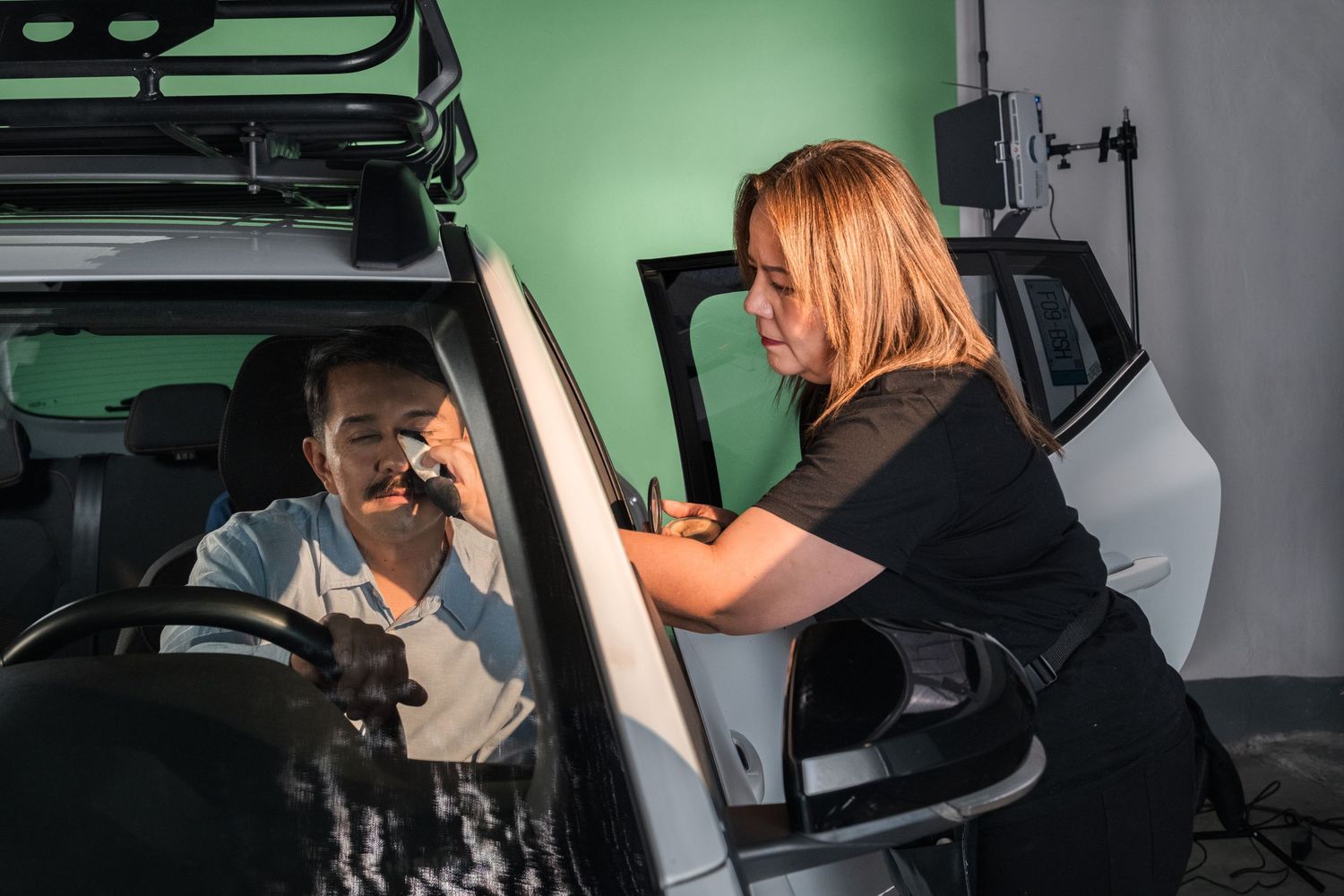 Makeup artist applying cosmetics to an actor sitting in a car on a film set.