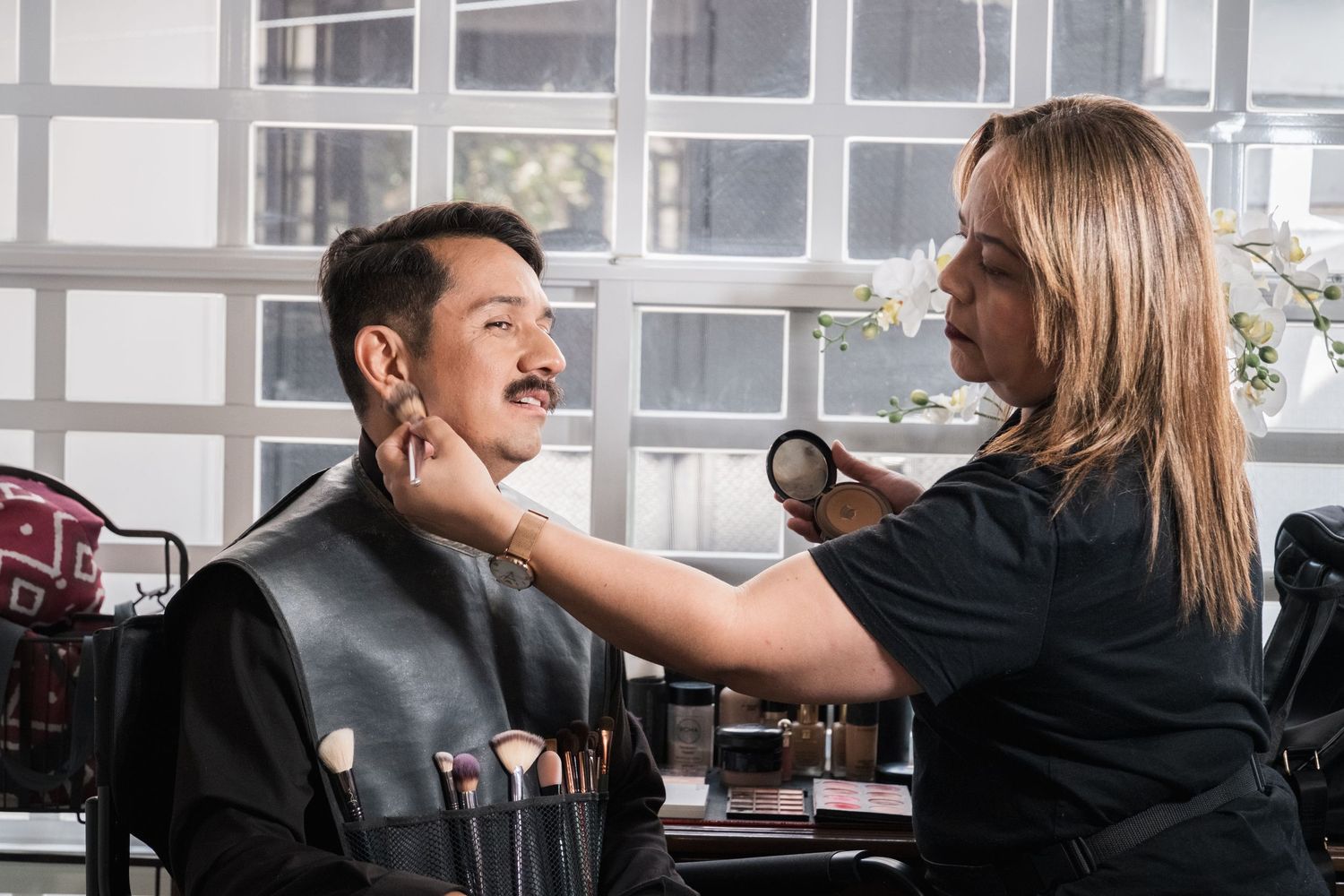 A makeup artist applies cosmetics to a man with a mustache in a professional makeup chair.