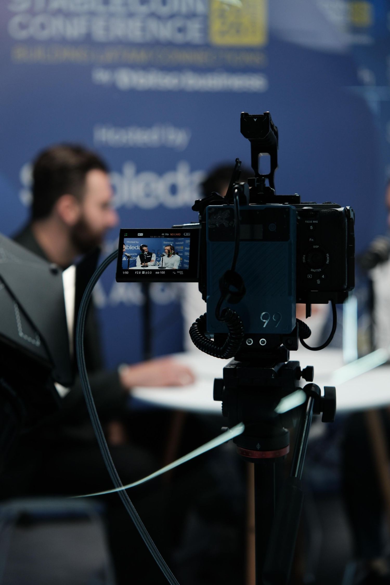 A professional camera on a tripod captures two speakers at the Stabledash Stablecoin Conference podcast studio.