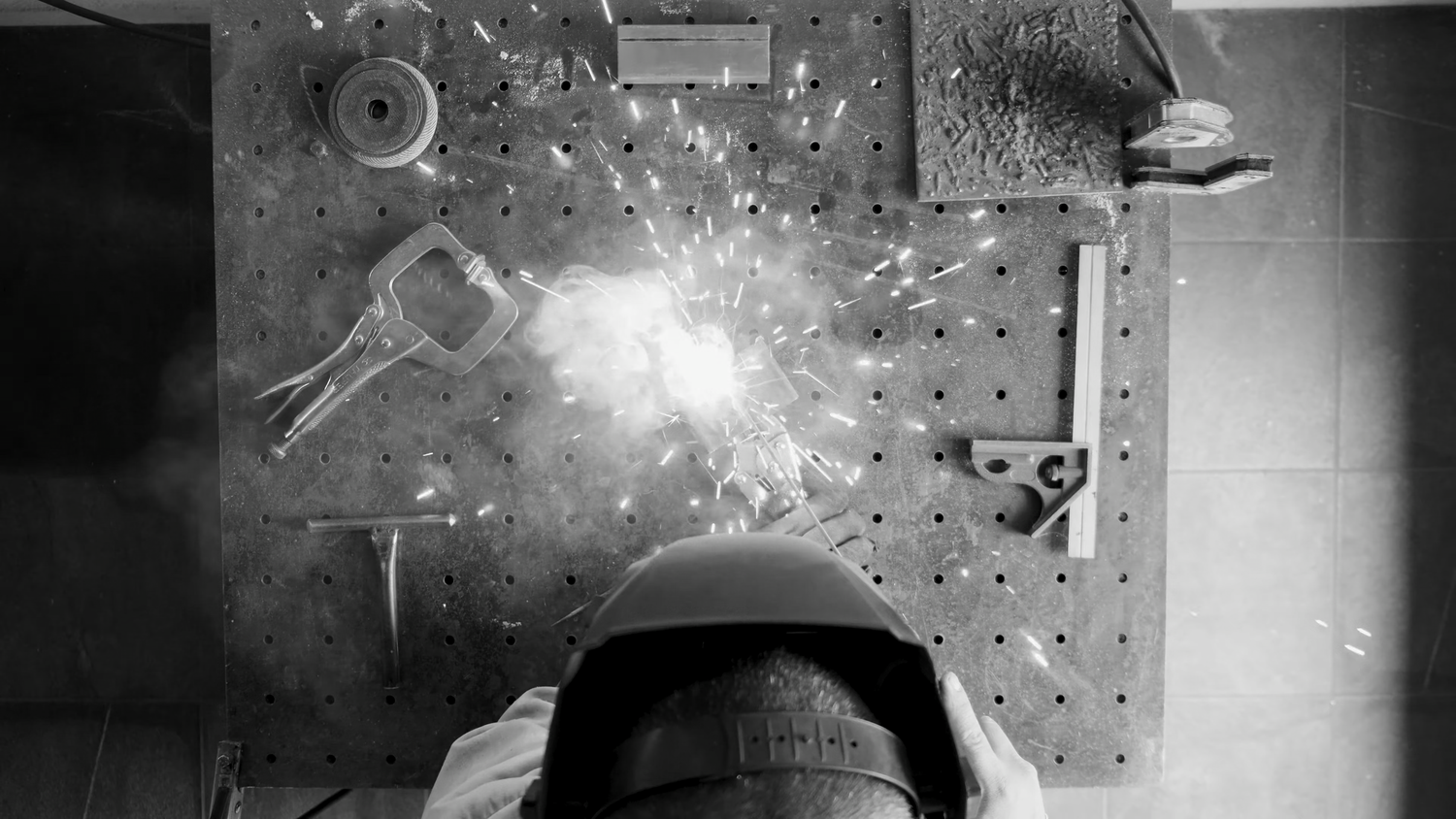 Overhead black and white shot of welder with sparks flying on perforated metal workbench.