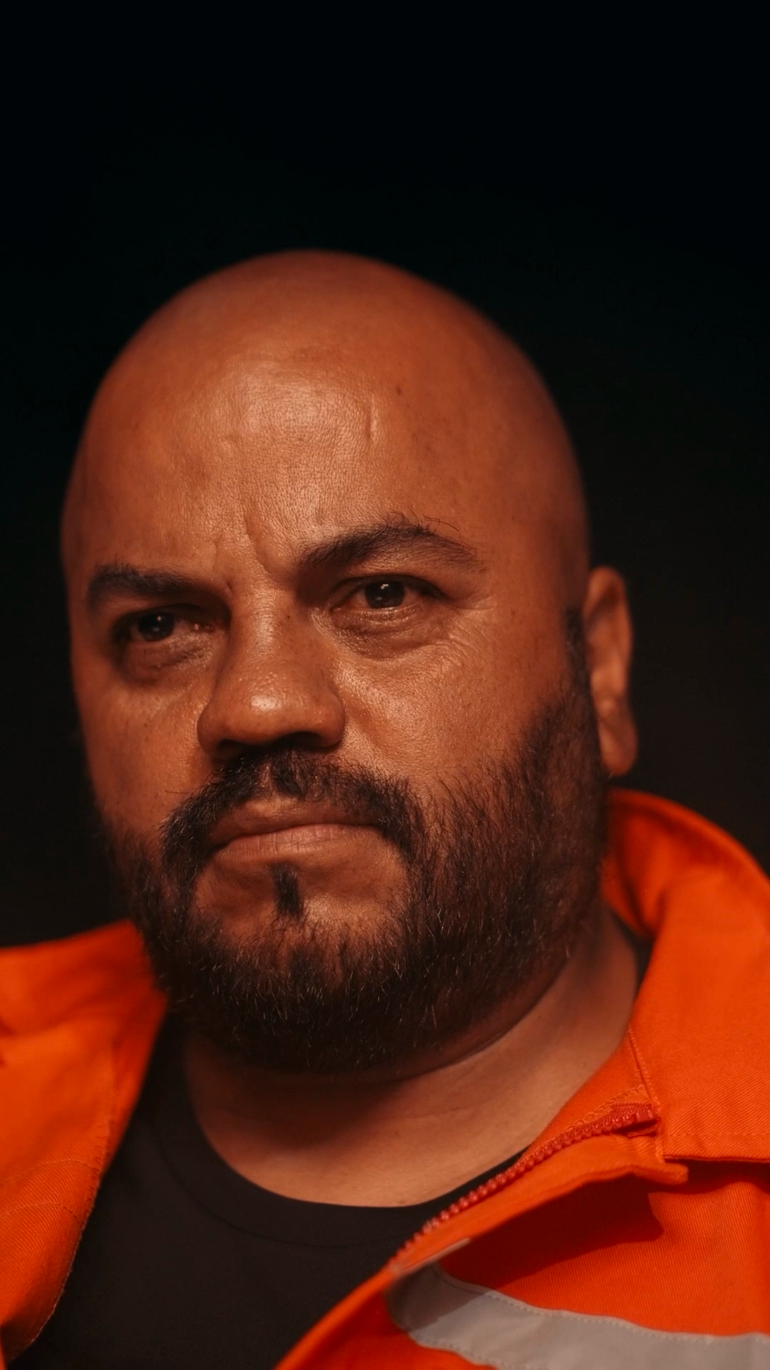 Bald man with beard wearing an orange jacket stares intensely against a dark background.