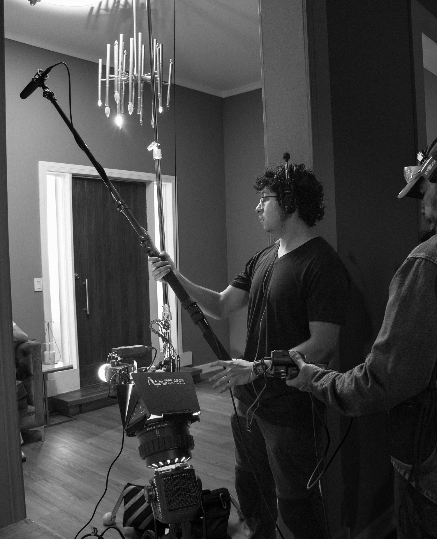 Black and white BTS photo of boom operator holding microphone pole next to Aputure light on set.