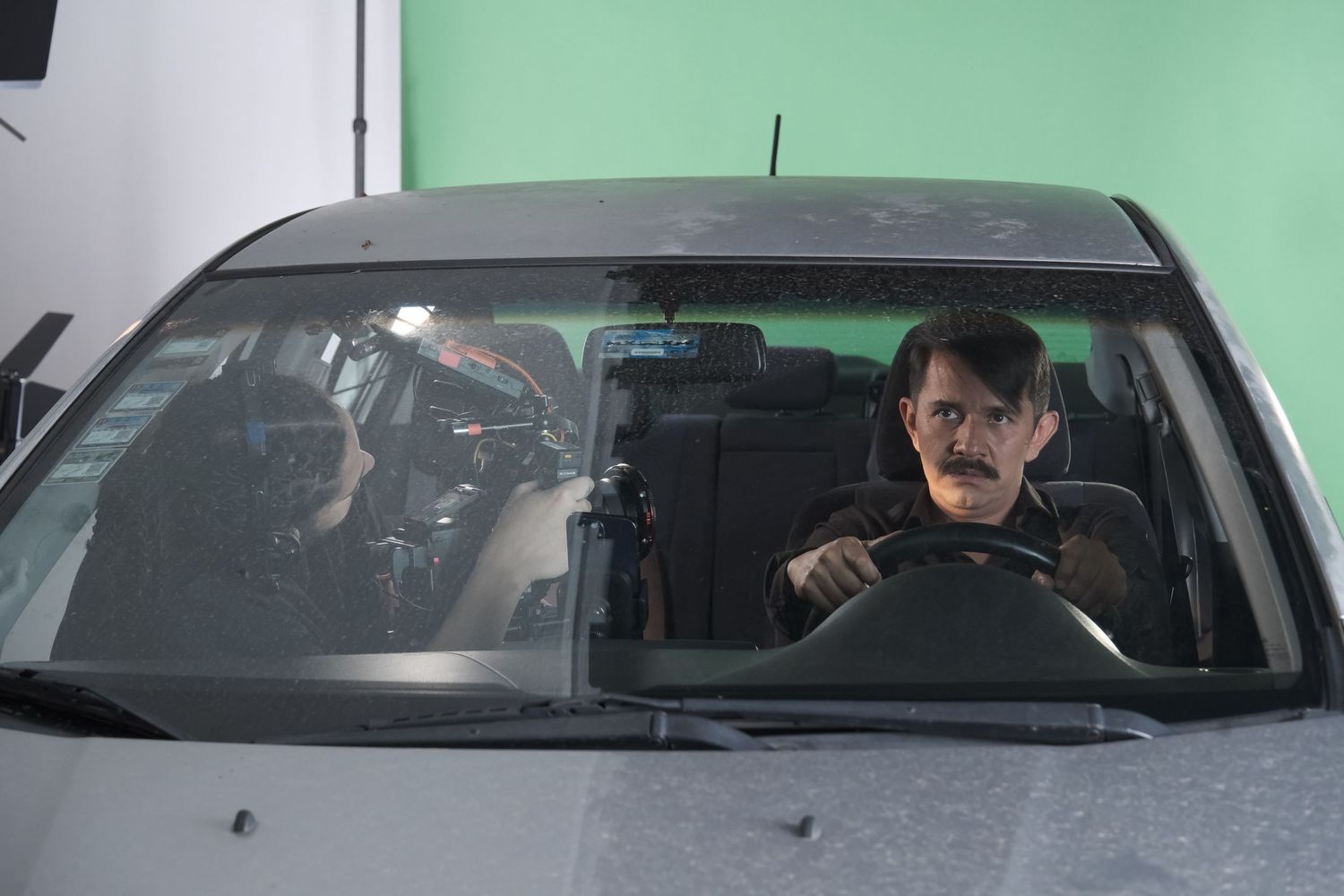 Film crew member operating a camera beside an actor with a mustache sitting in a car on green screen.