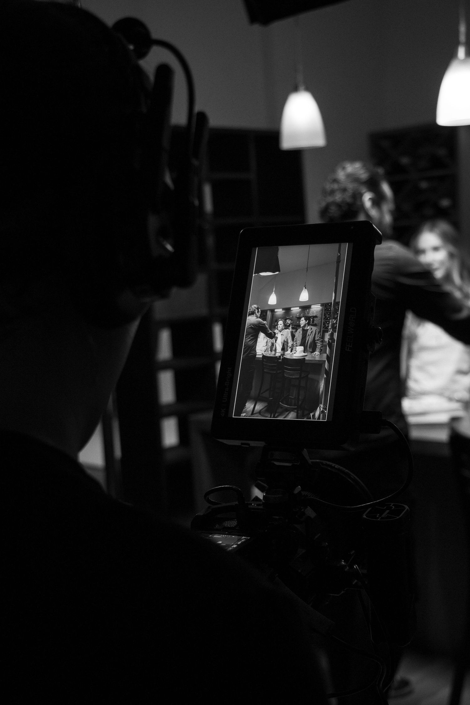 Behind-the-scenes black and white shot of a cameraman filming a group scene through a monitor on set.