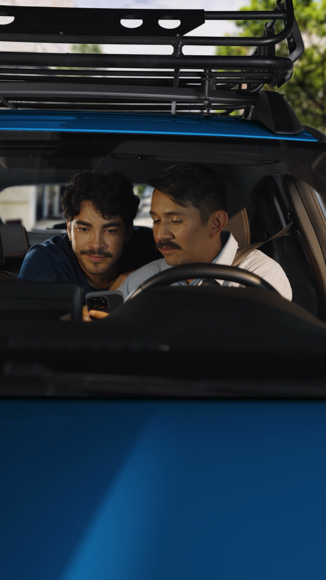 Two men sitting inside a blue SUV looking at a smartphone together during the day.