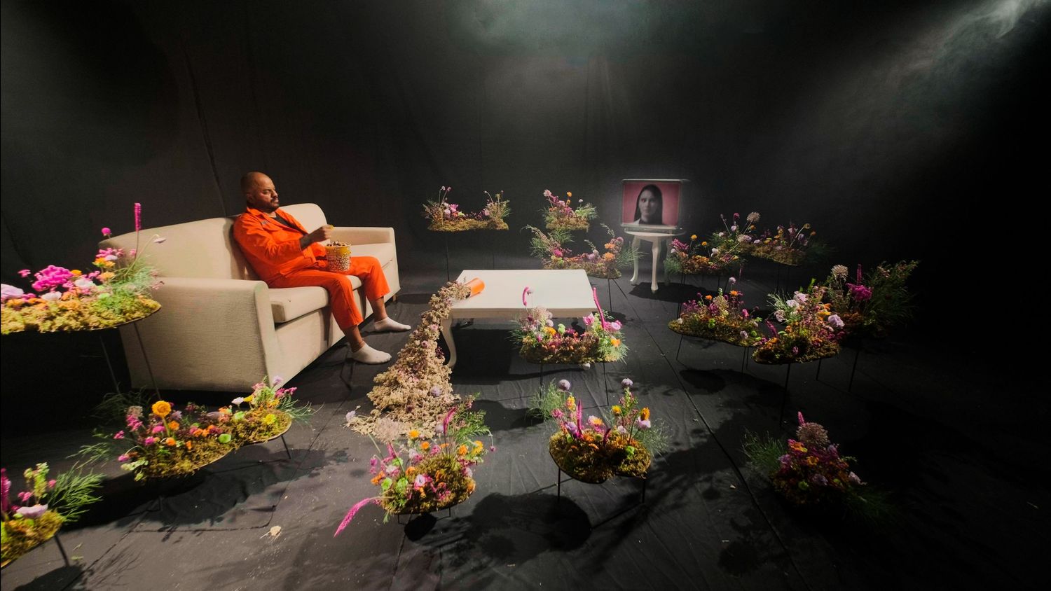 Person in orange jumpsuit sitting on white sofa surrounded by colorful flowers in a dark studio set.